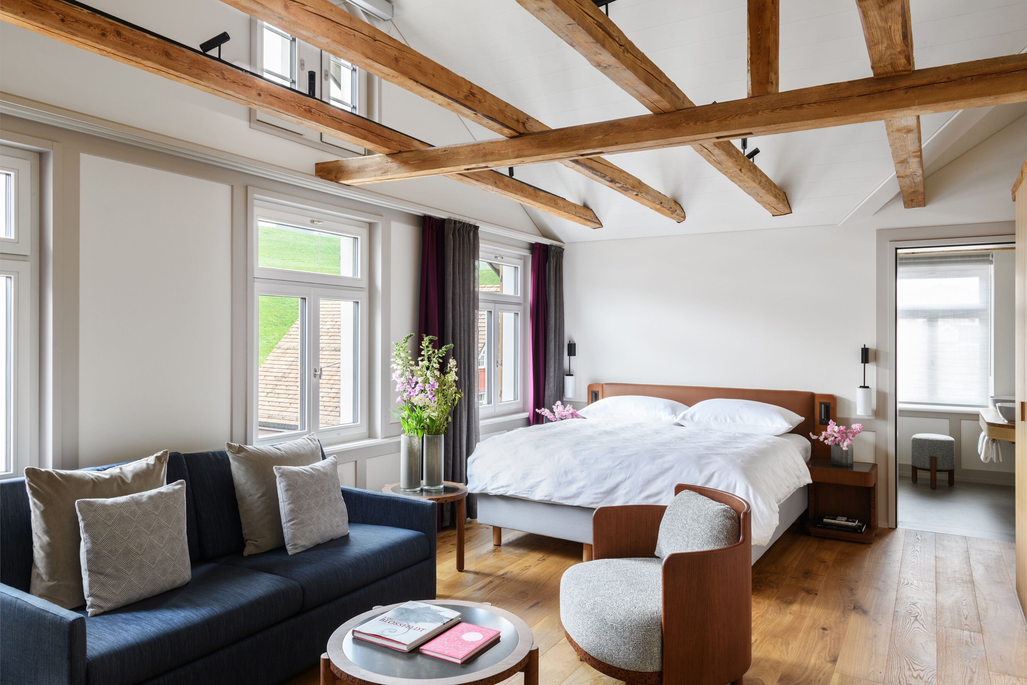 Un dormitorio moderno con una cama grande, un sofá, una silla y una mesita pequeña. Hay ventanas con cortinas, techo de madera y vistas al exterior con vegetación.