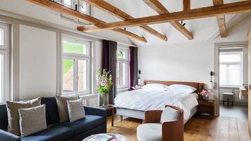 Un dormitorio moderno con una cama grande, un sofá, una silla y una mesita pequeña. Hay ventanas con cortinas, techo de madera y vistas al exterior con vegetación.