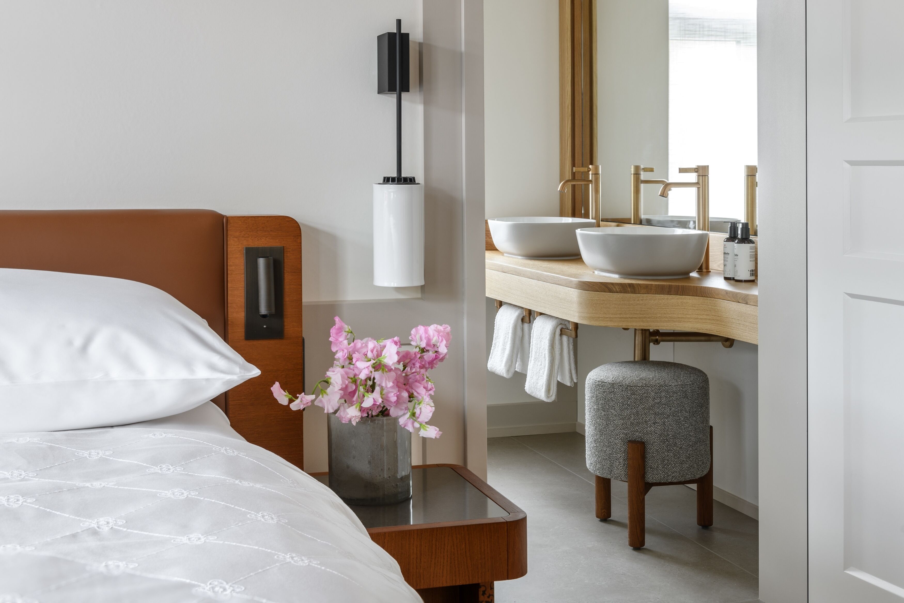 Een hotelkamer met een bed, een nachtkastje met een bloevas, een moderne badkamer met een wastafel en spiegel, en een houten kruk.