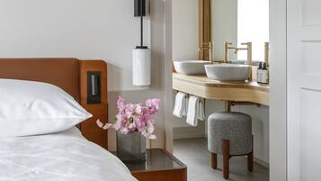 Een hotelkamer met een bed, een nachtkastje met een bloevas, een moderne badkamer met een wastafel en spiegel, en een houten kruk.