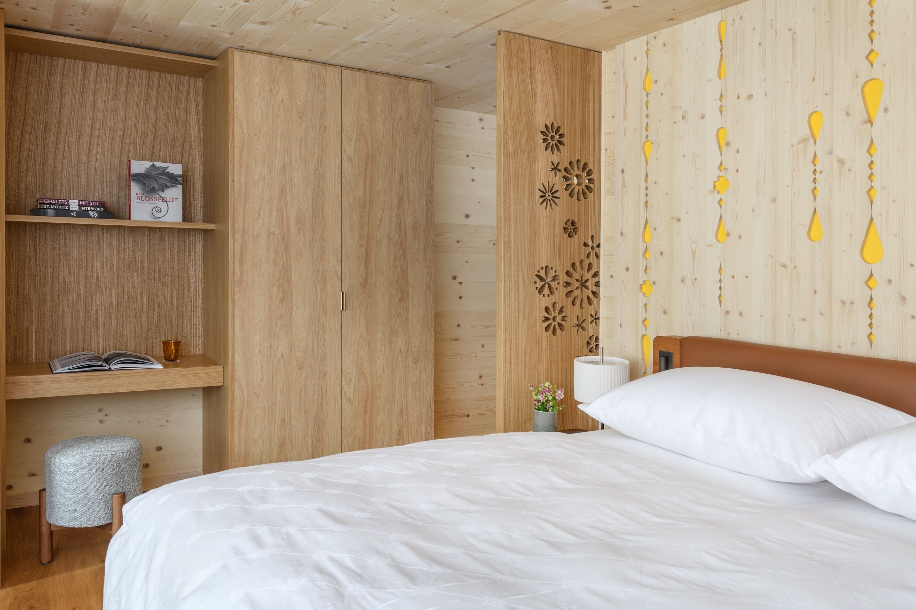 A bedroom with a wooden wardrobe, a bedside table with a lamp, and a bed with white bedding.