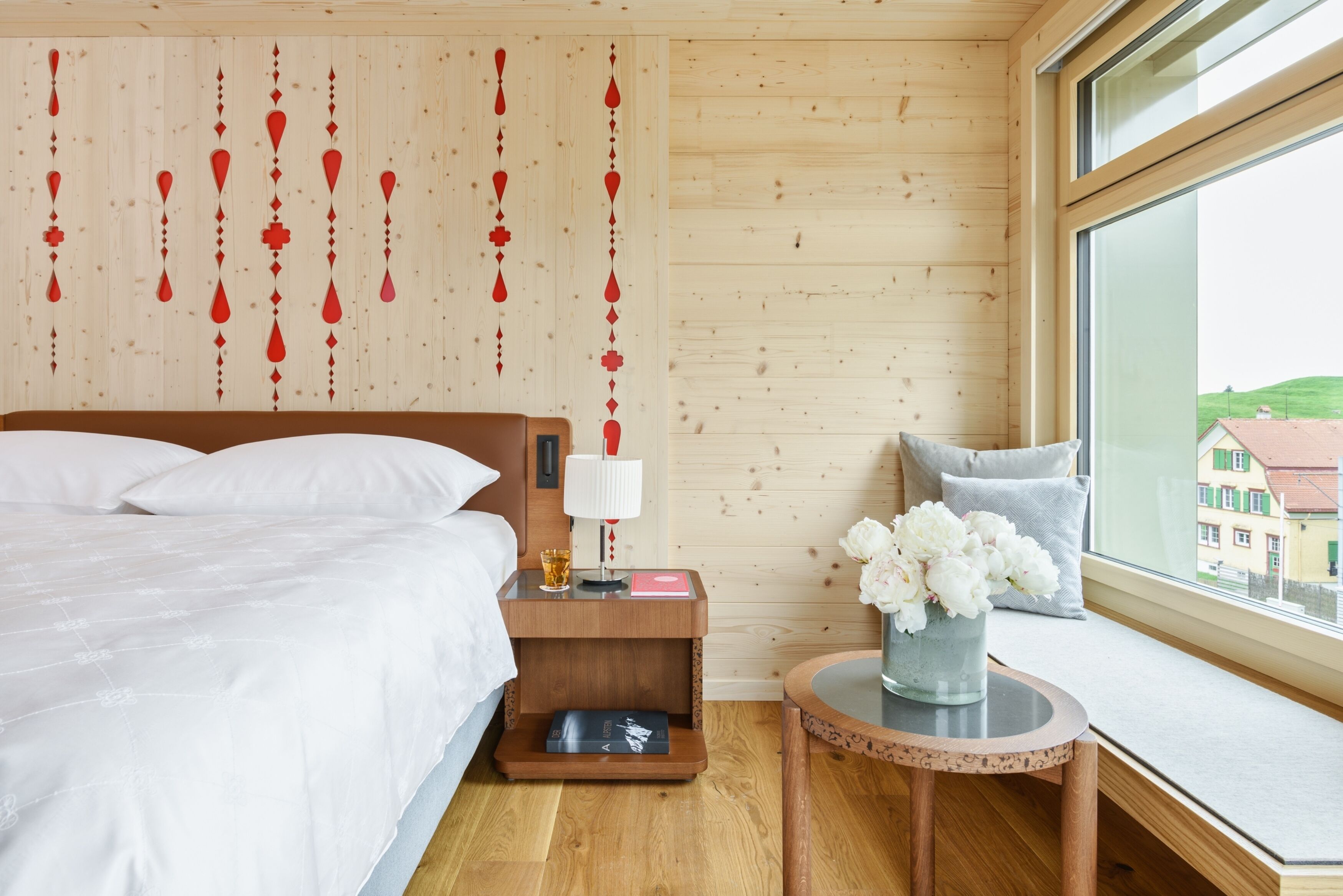 A bedroom with a wooden headboard, a bedside table with a lamp, a small round table with a vase of flowers, and a window with a view of buildings.