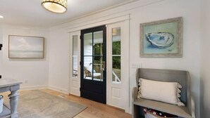 Un couloir doté d’un plancher en bois, d’une porte noire avec des panneaux de verre, d’un mur blanc orné d’un tableau encadré et d’un banc avec un coussin.