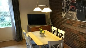 A dining area with a wooden table and white chairs, a television on a stand, and a brick wall with a street scene painting. - Vacation home directly on the dike Pet friendly (Wurster Nordseeküste)