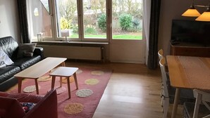 A living room with a sofa, coffee table, and TV. There are large windows with curtains, a radiator, and a view of greenery outside. - Vacation home directly on the dike Pet friendly (Wurster Nordseeküste)