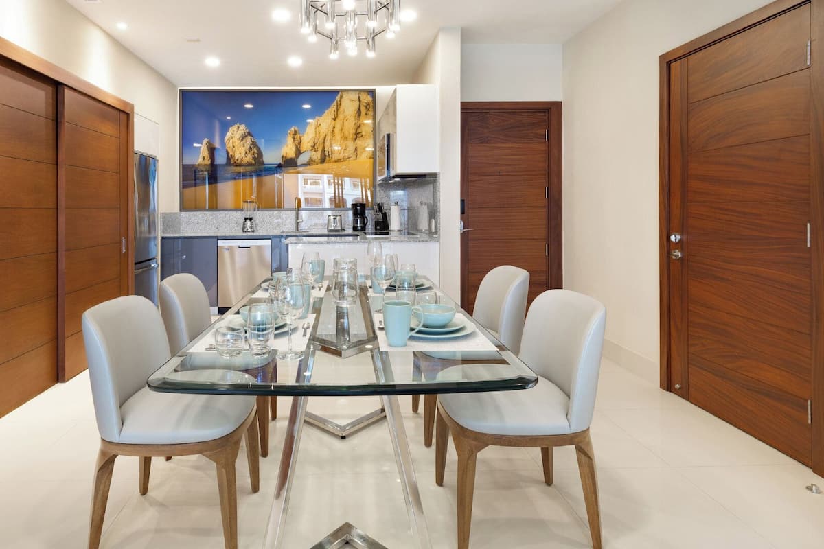 A dining area with a glass table, white chairs, and a kitchen in the background.