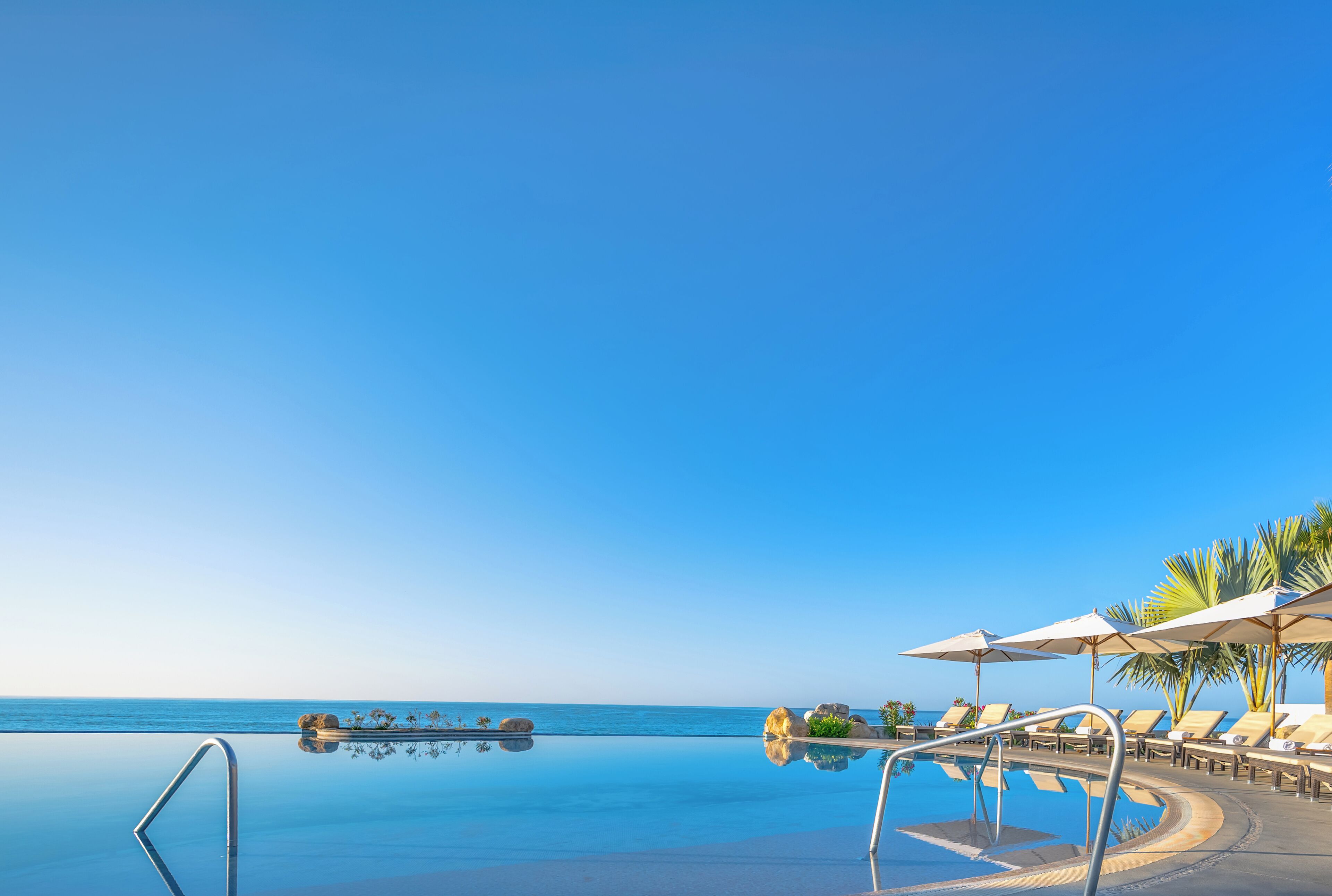 A clear blue sky, an infinity pool, and lounge chairs with umbrellas.
