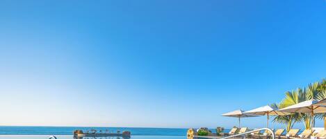 A clear blue sky, an infinity pool, and lounge chairs with umbrellas.
