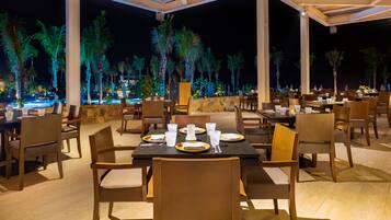 An outdoor dining area with tables and chairs, palm trees, and a pool in the background.