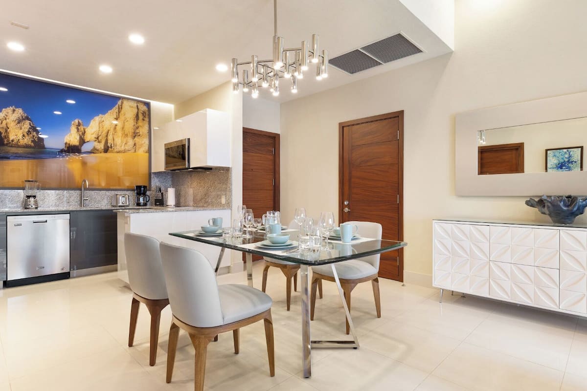 A modern kitchen with a dining area, featuring a glass table, chairs, and a decorative wall piece.