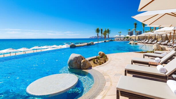 Zona de piscina con sillas de descanso y sombrillas, cielo azul despejado y vista al océano.