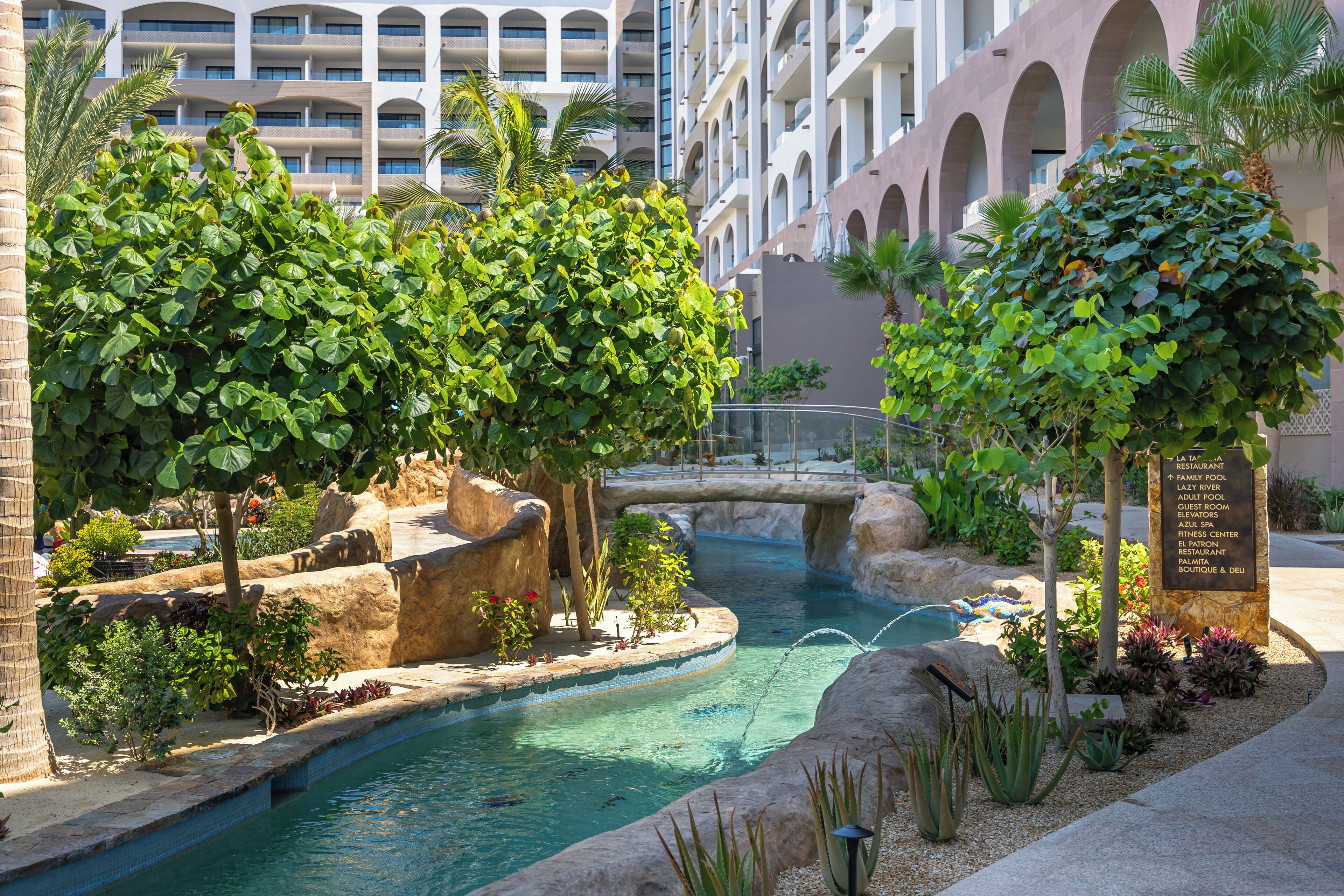 A hotel pool area with a water feature, surrounded by greenery and a multi-story building in the background.
