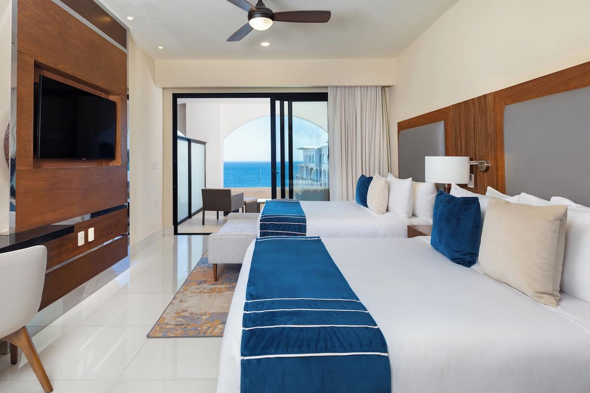 A hotel room with two beds, a TV, a ceiling fan, and a view of the sea.