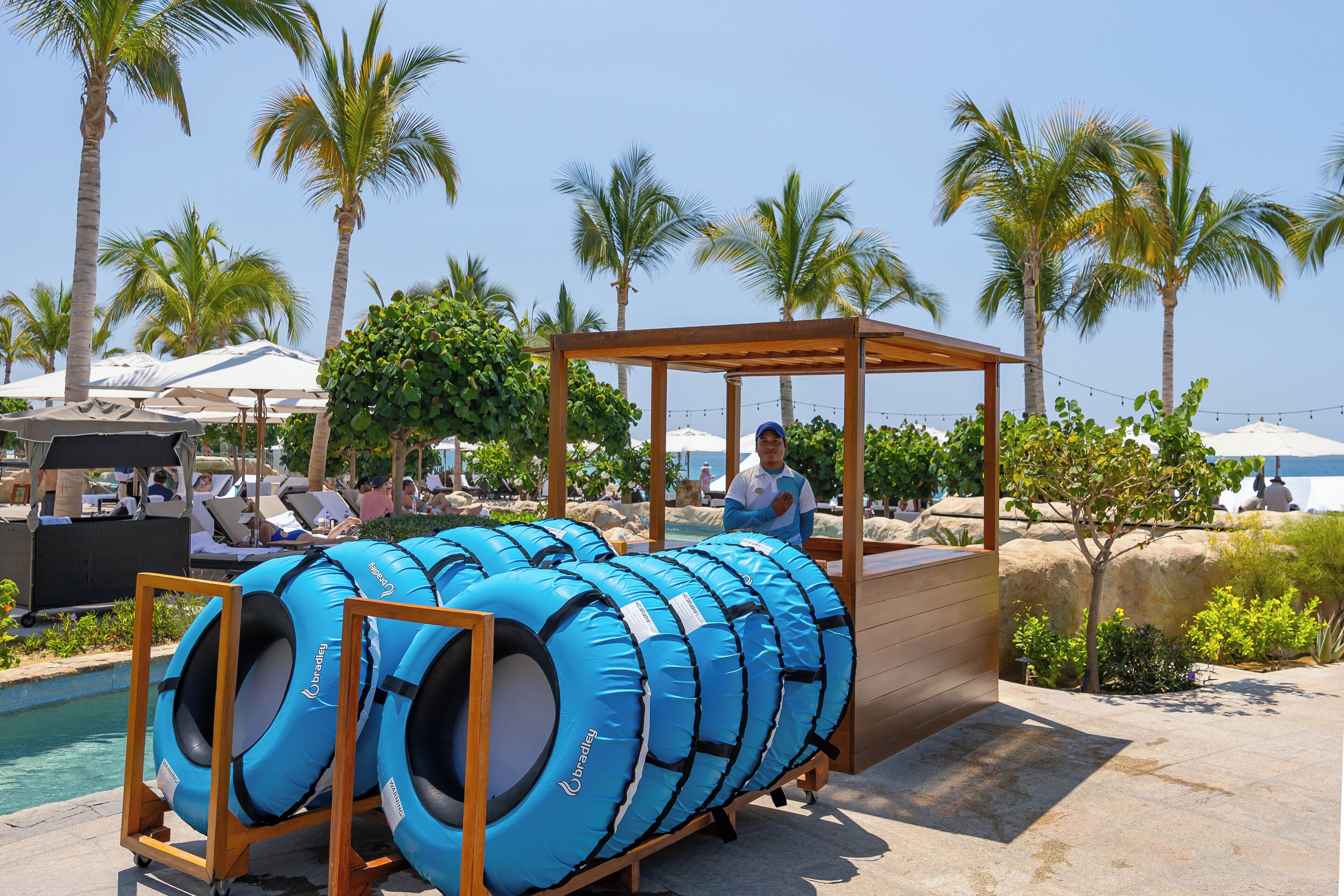 Un espace aménagé au bord de la piscine, avec des chaises longues et des parasols, une pergola en bois et des bouées gonflables bleues.