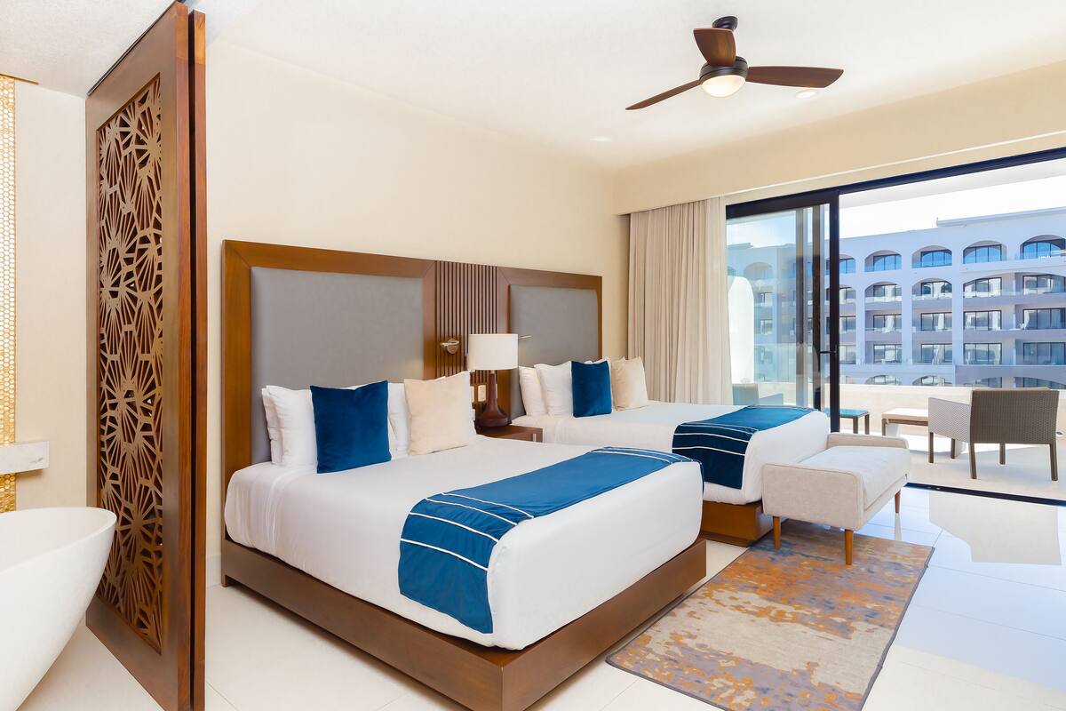 A hotel room with two beds, a large window with a view of buildings, a ceiling fan, and a modern bathroom.