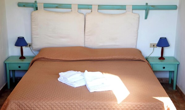 Un lit recouvert d’une couette marron, avec deux oreillers et une pile de serviettes blanches posées sur une tête de lit de couleur turquoise. Deux tables de chevet, chacune surmontée d’une lampe, se trouvent de part et d’autre du lit.