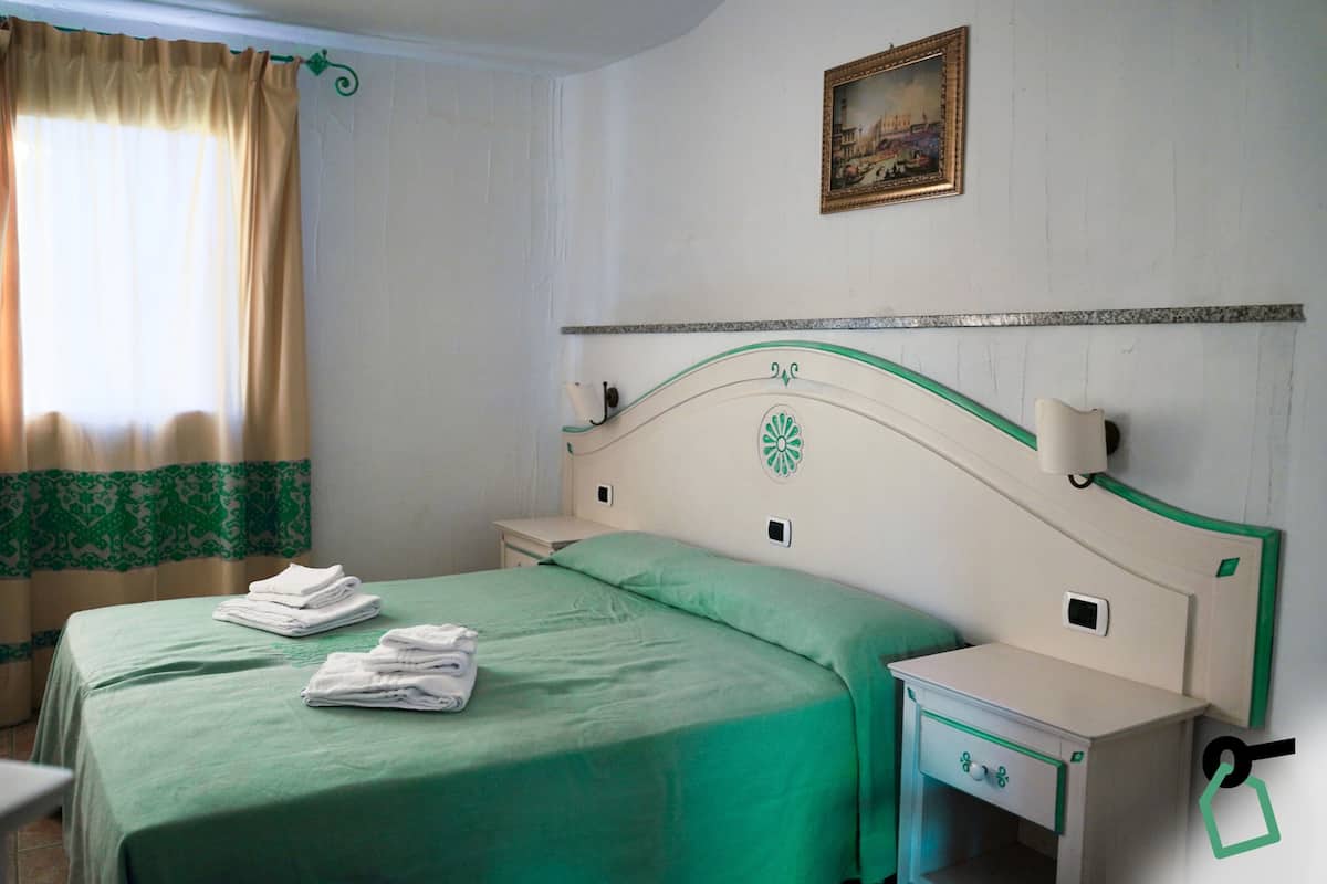 A neatly made bed with green bedding and white towels, a small bedside table with a drawer, and a painting on the wall.