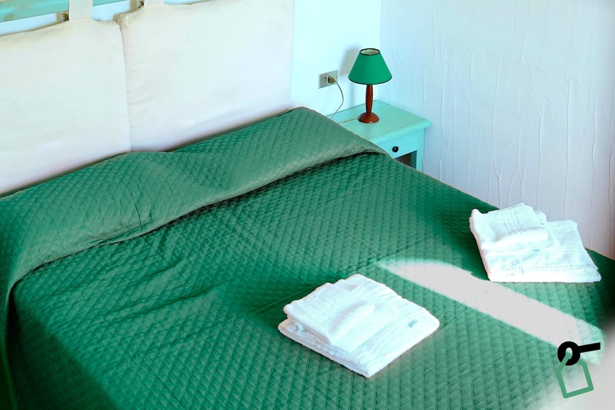 A bed with a green quilt, a green lamp on a nightstand, and two folded towels on the bed.