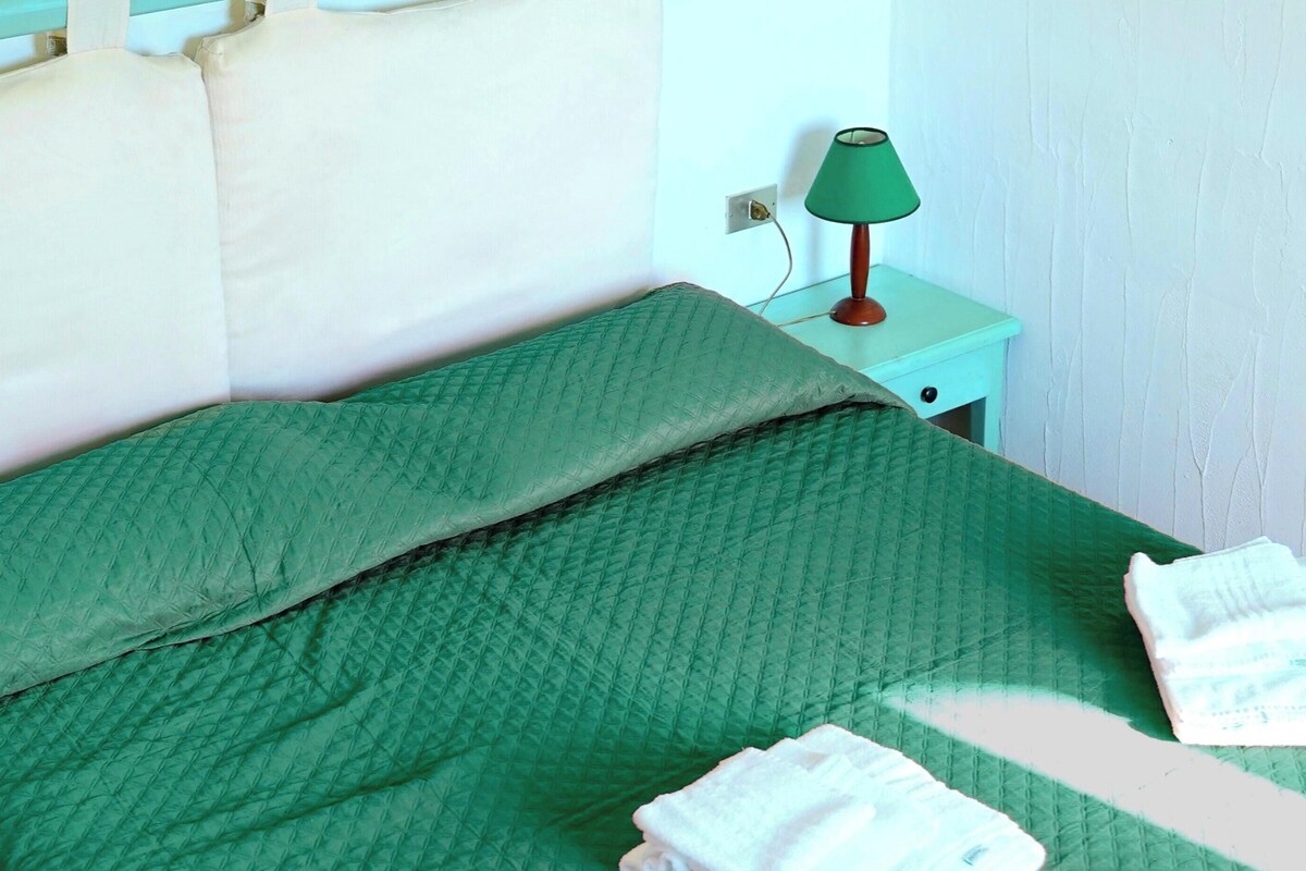 Un lit recouvert d’une couette verte, une table de chevet avec une lampe verte et un mur blanc.