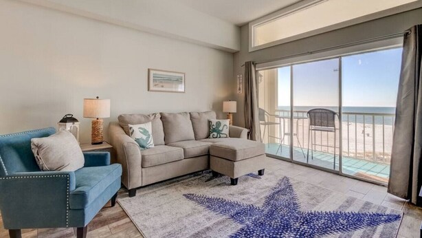 A living room with a sofa, a chair, a coffee table, and a large window with a view of the beach.