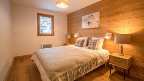 Une chambre à coucher avec une tête de lit en bois, un lit recouvert d’une couette blanche, deux tables de chevet avec des lampes et une fenêtre donnant sur des immeubles.