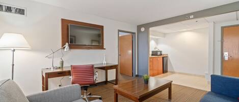 A modern living room with a sofa, a coffee table, a desk with a lamp, a television, and a kitchenette with a microwave.