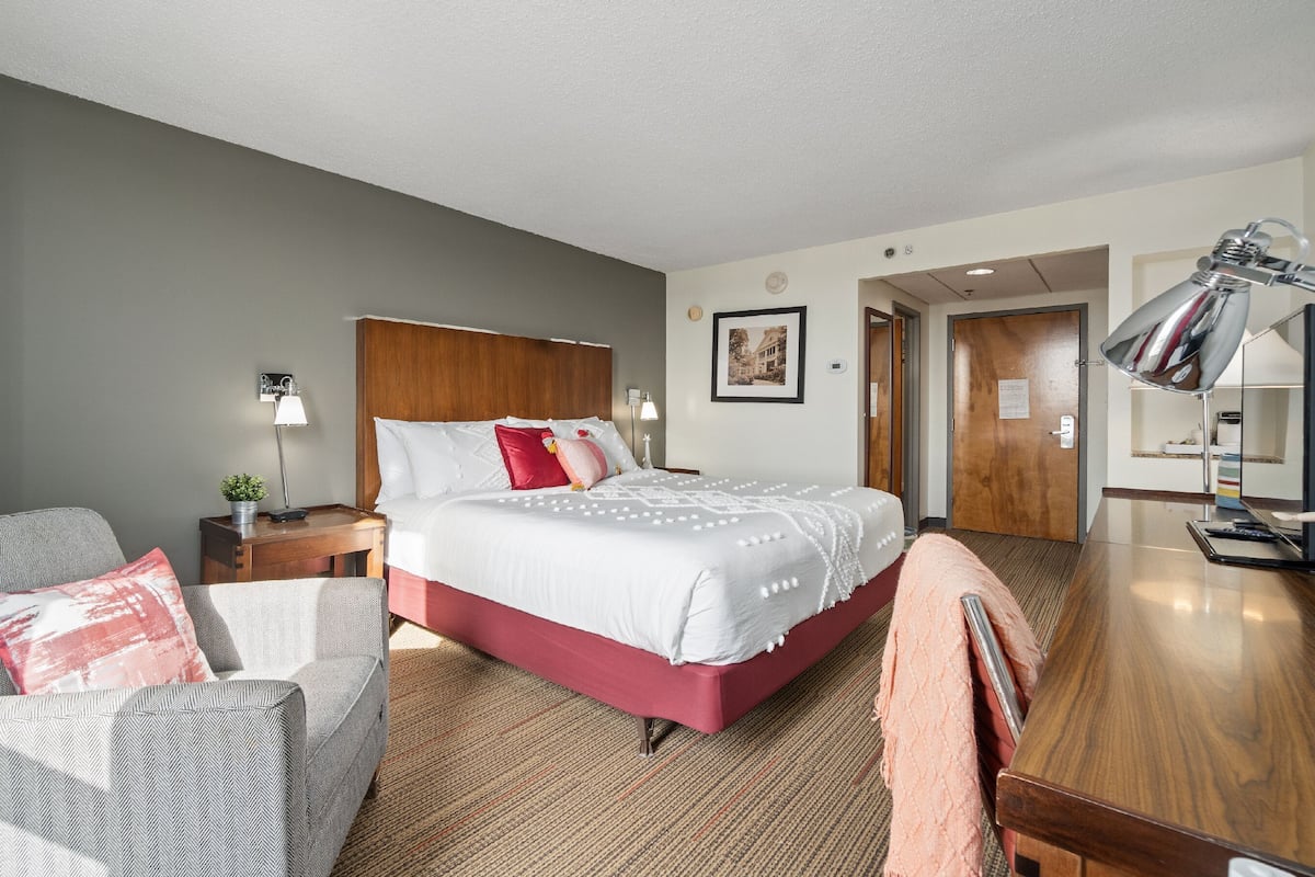 A hotel room with a bed, a desk, a chair, a lamp, a nightstand, and a door.