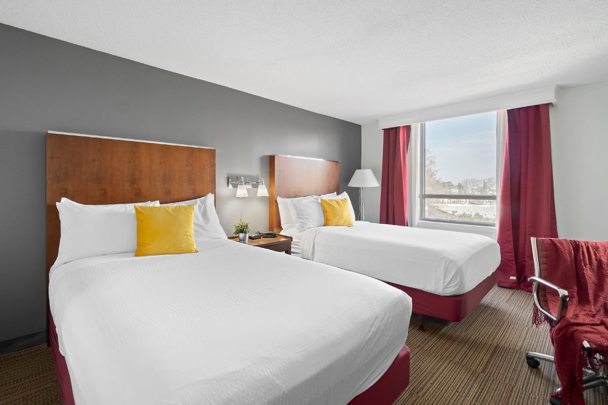 A hotel room with two beds, a wooden headboard, a nightstand with a lamp, a window with curtains, and a view of the outdoors.