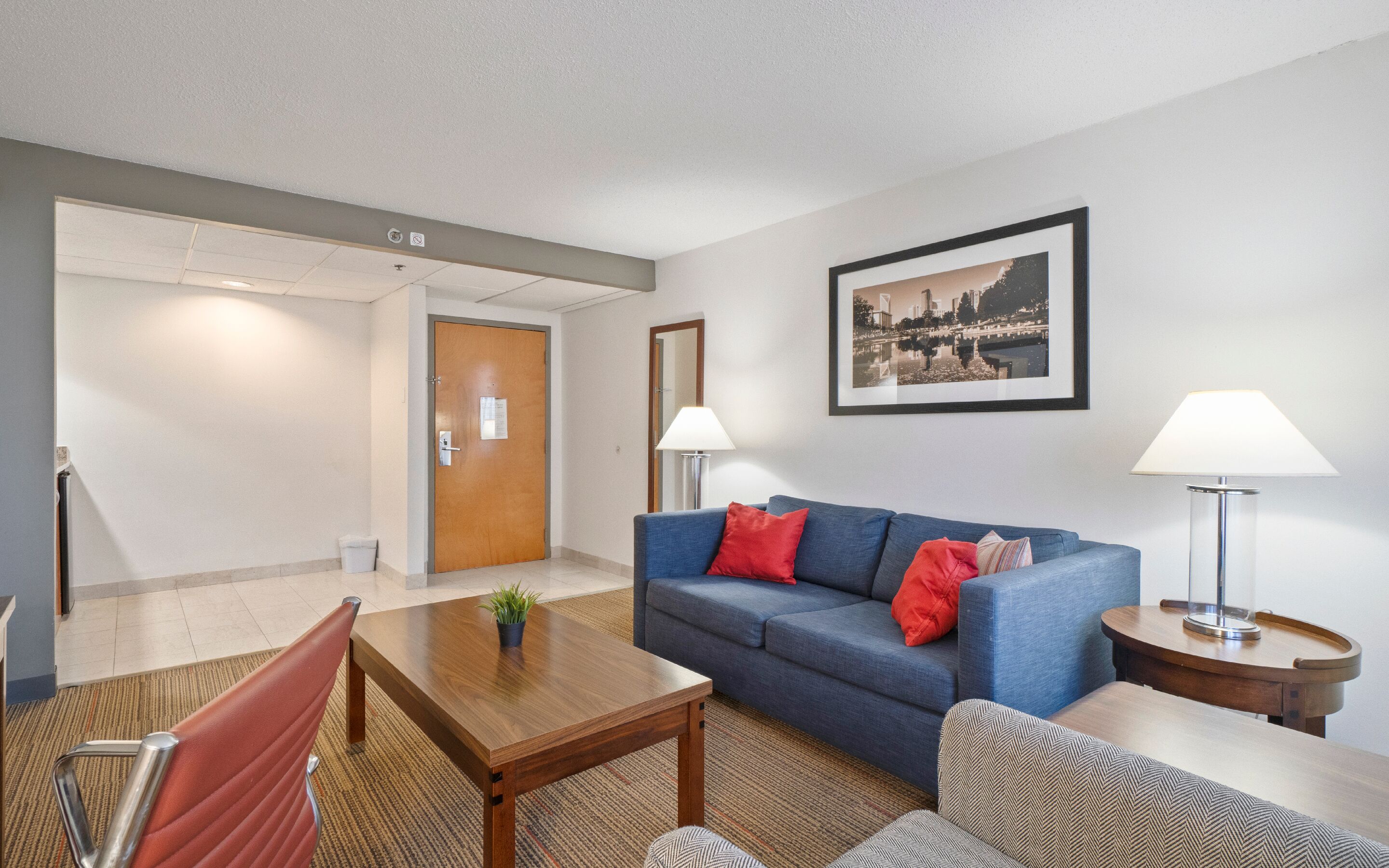 Een moderne hotelkamer met een blauwe bank, rode kussens, een houten salontafel en een lijst met een stadsbeeld aan de muur.