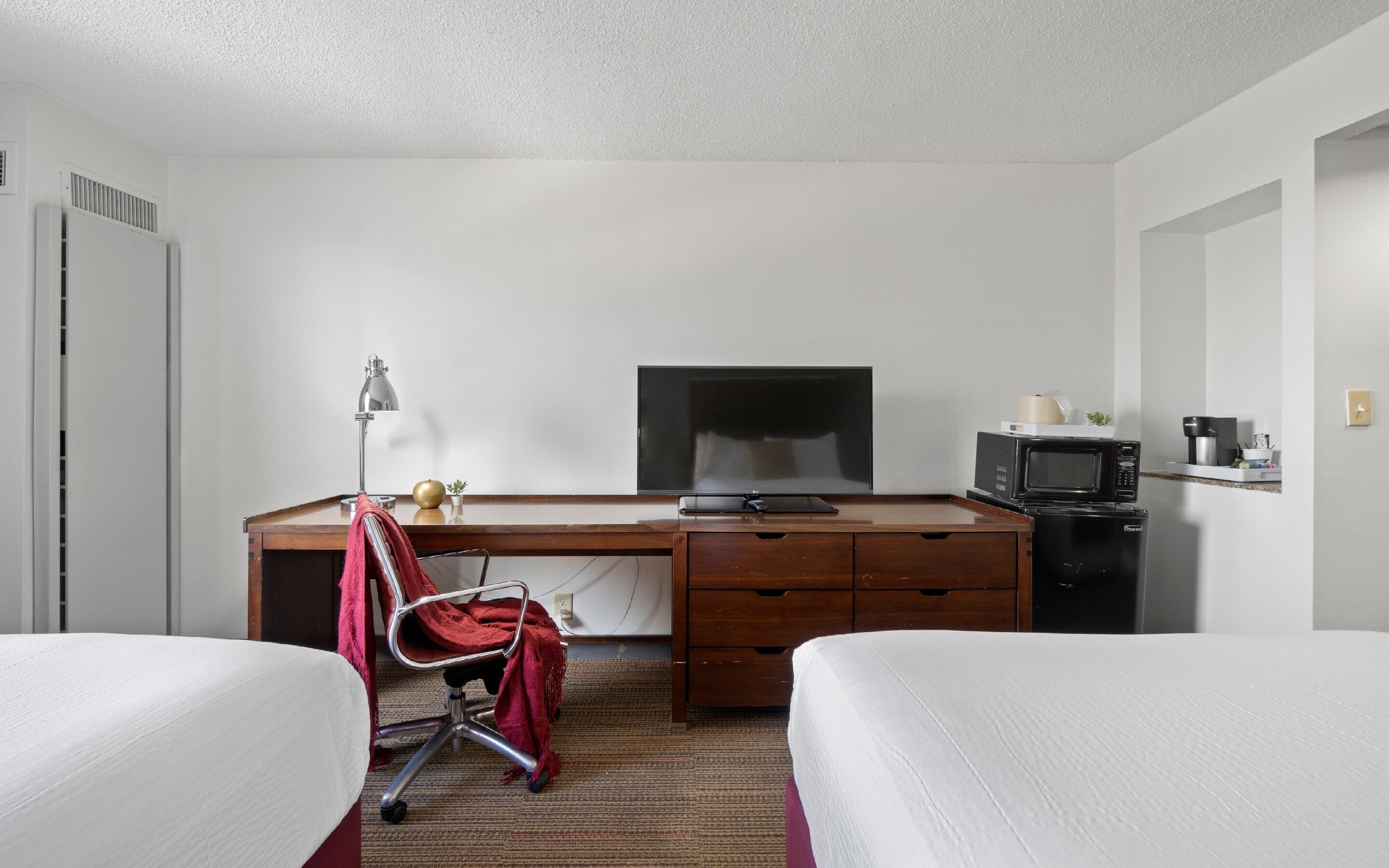 Habitación de hotel con dos camas, un escritorio con televisor, microondas y una silla con una manta roja.