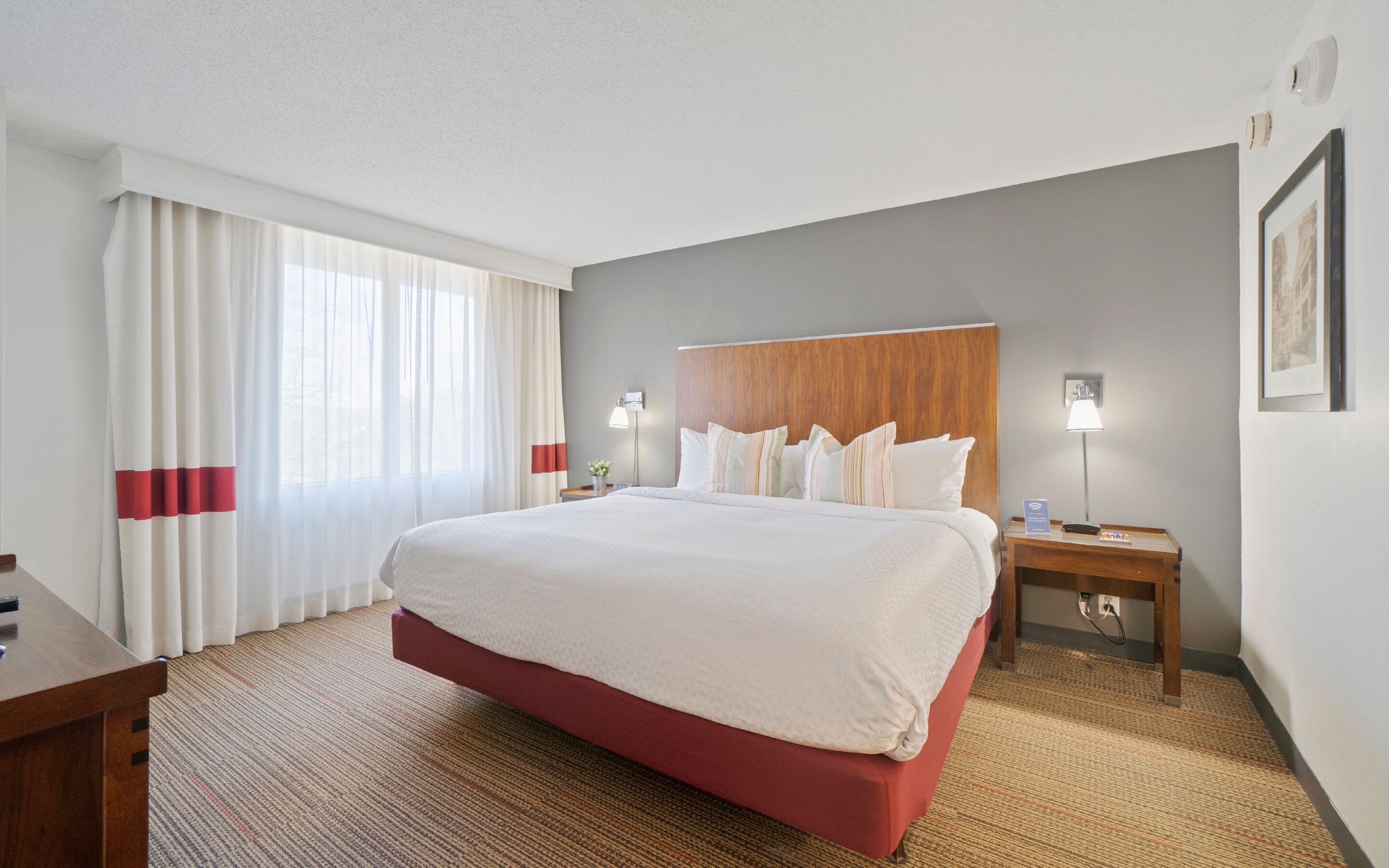 A hotel room with a large bed, a wooden headboard, two bedside tables with lamps, a desk, and a window with curtains.