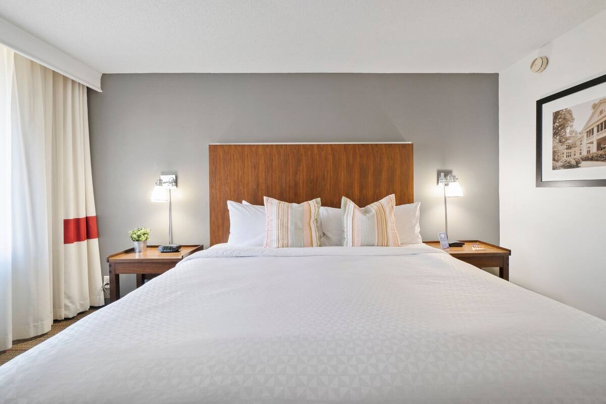 A hotel room with a large bed, two bedside tables, a wooden headboard, a wall-mounted picture, and a window with curtains.