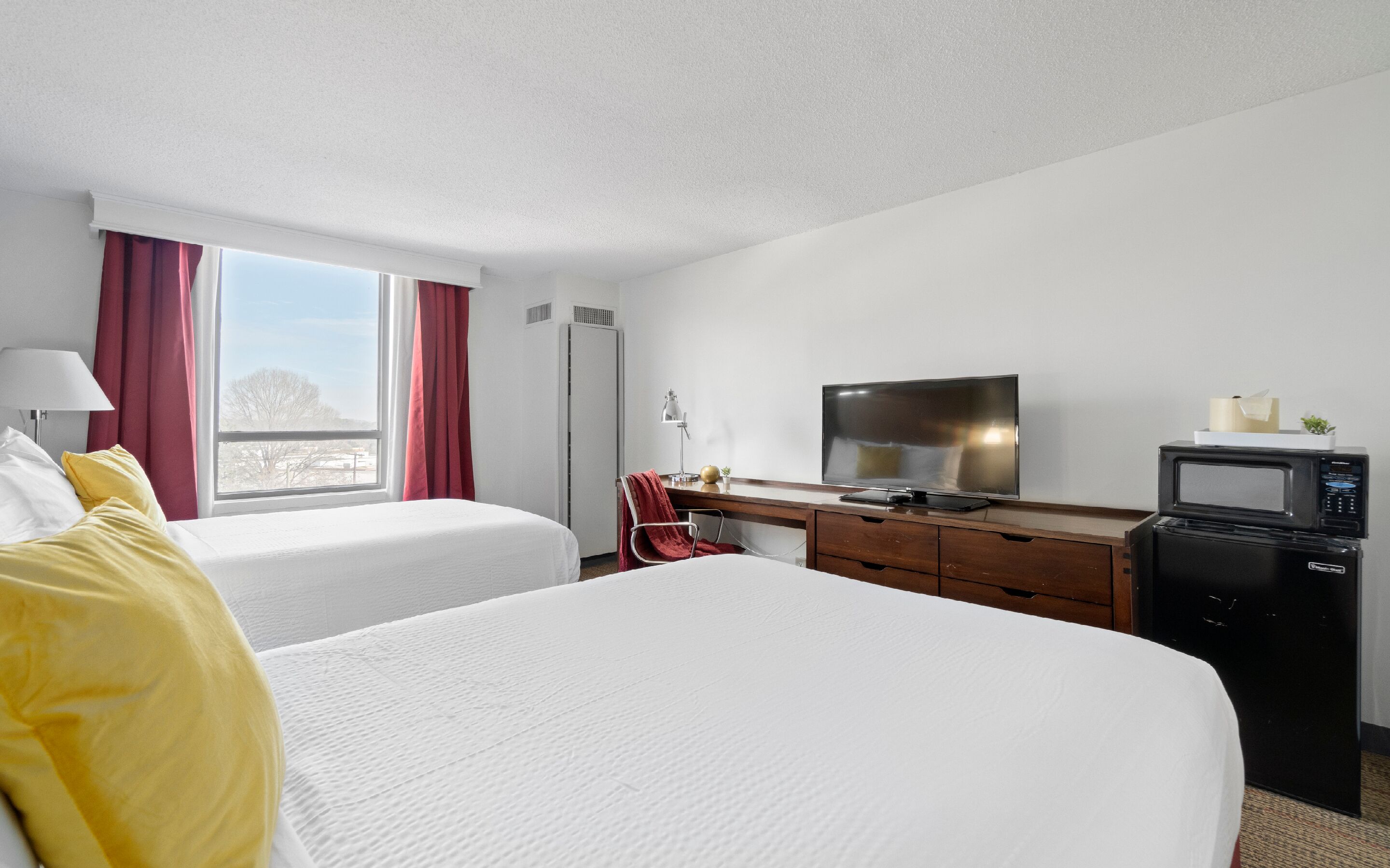 Habitación de hotel con dos camas, un escritorio con televisor y microondas, una ventana con cortinas rojas y una lámpara.