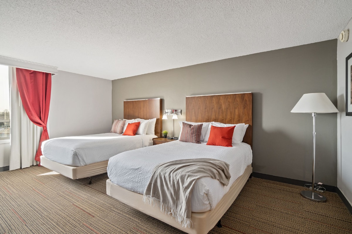 A hotel room with two beds, a large window with red curtains, a floor lamp, and a framed picture on the wall.