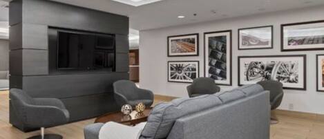 Una sala moderna con un sofá gris, dos sillones, una mesa de centro y un televisor de pantalla plana montado en una pared oscura.