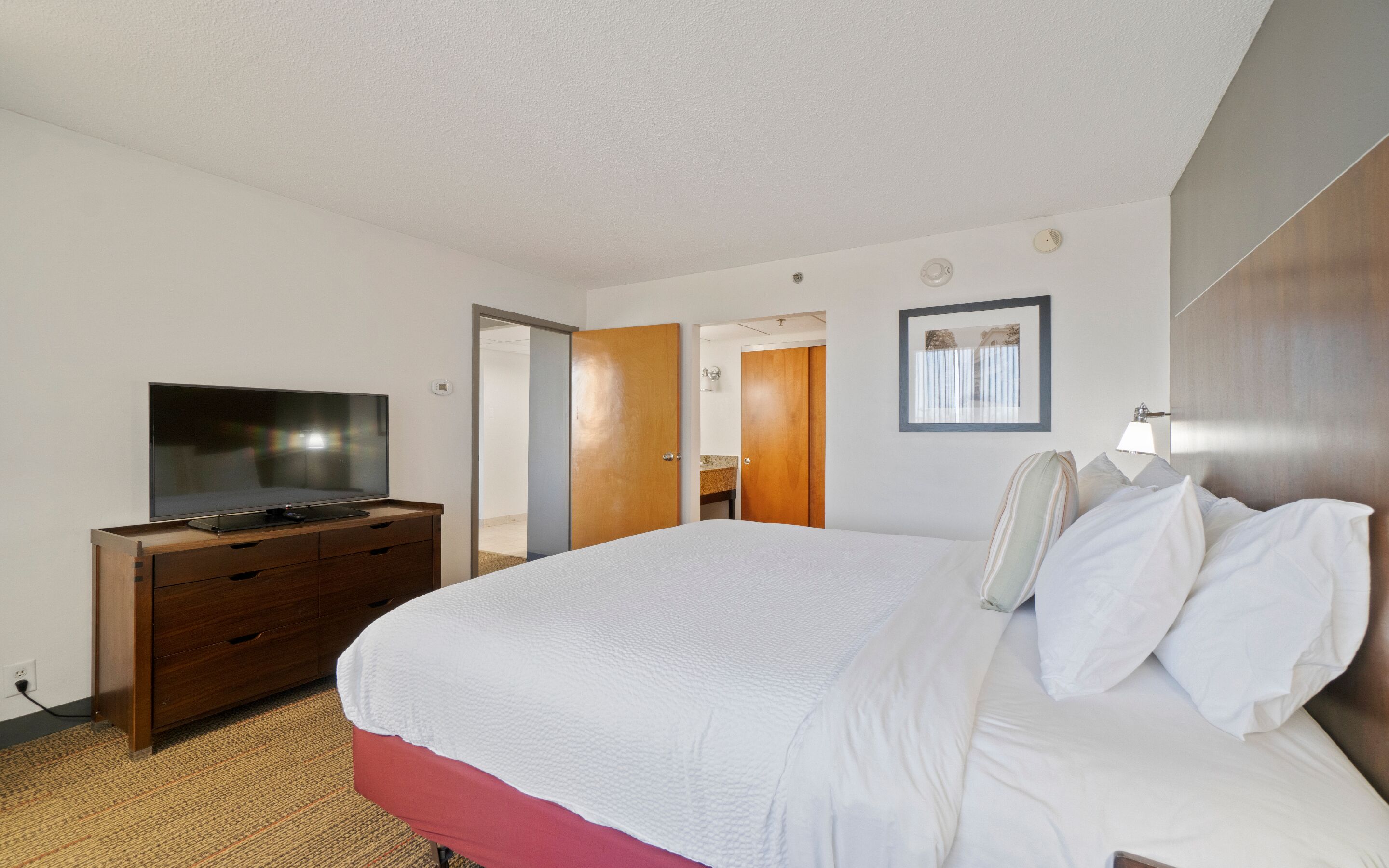A hotel room with a bed, a TV on a wooden dresser, a bathroom visible through an open door, and a framed picture on the wall.