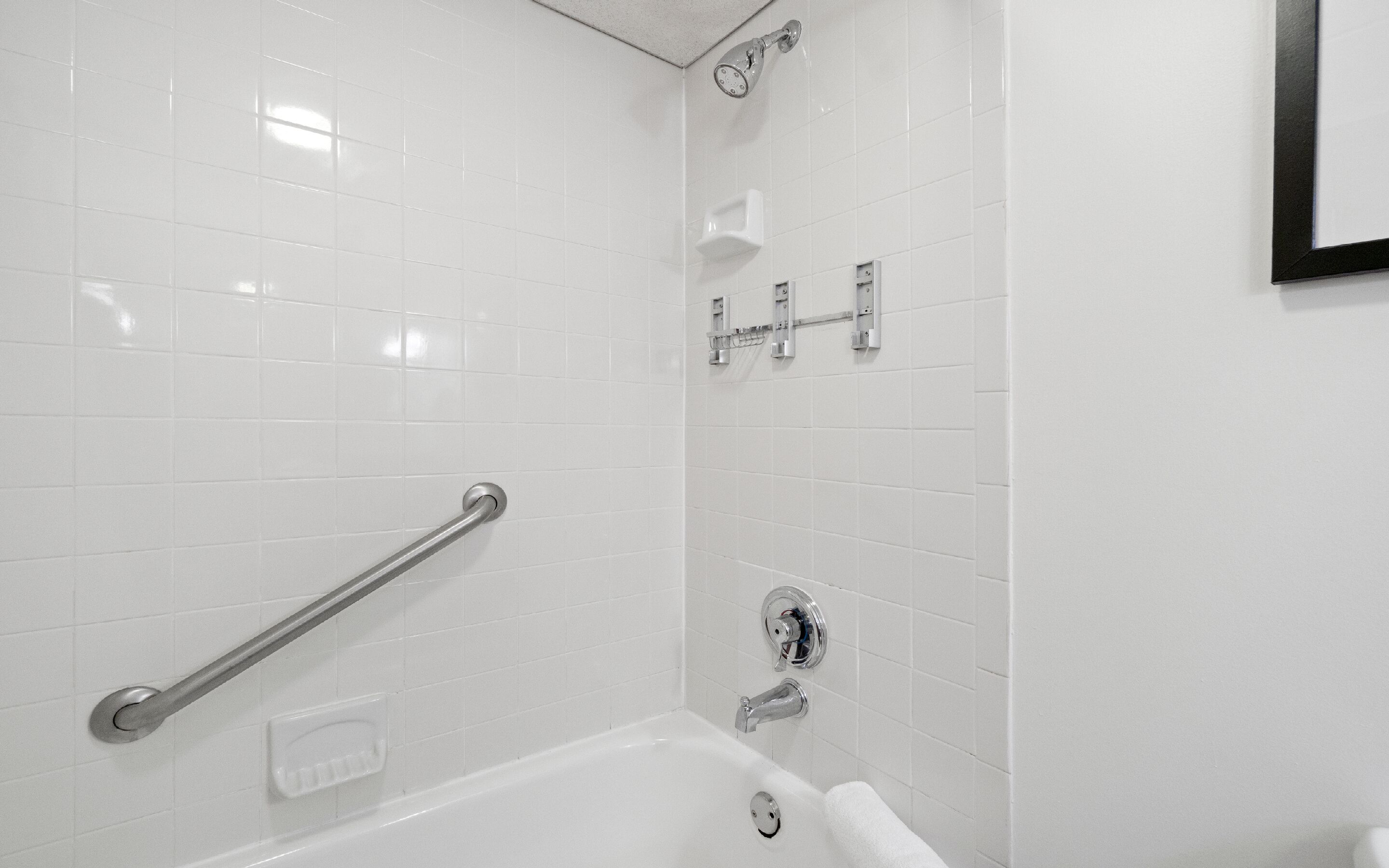 A bathroom with white tiled walls, a bathtub, a shower with a handheld showerhead, and a grab bar.