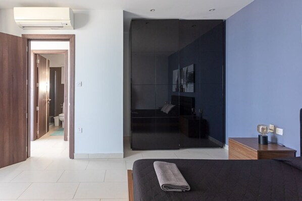 A modern bedroom with a bed, a nightstand, a mirror, and a sliding door leading to a bathroom. - Sea-View Apartment at WatersEdge Complex with two pools (Marsaskala)