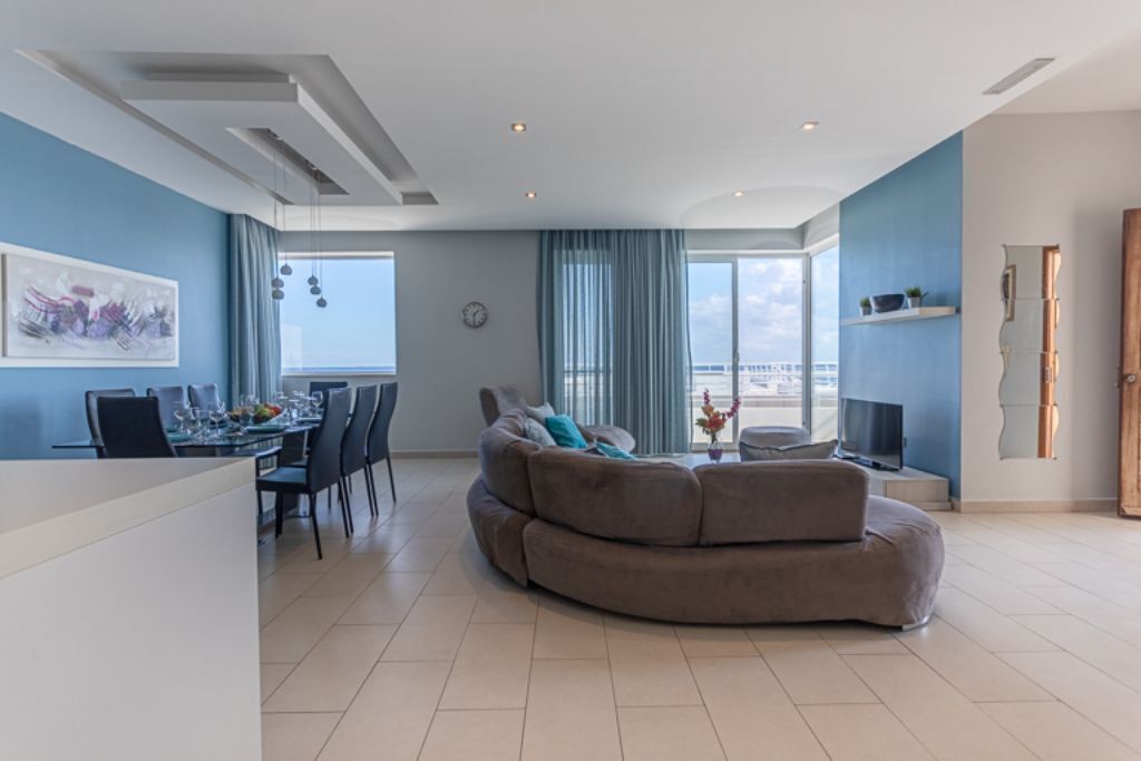 Un salon moderne avec un canapé aux lignes courbes, un coin repas avec une table dressée pour quatre personnes et une grande fenêtre offrant une vue sur la mer.
