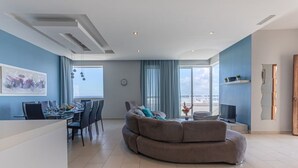 A modern living room with a curved sofa, a dining area with a table set for four, and a large window offering a view of the sea. - Sea-View Apartment at WatersEdge Complex with two pools (Marsaskala)
