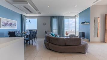Un salon moderne avec un canapé aux lignes courbes, un coin repas avec une table dressée pour quatre personnes et une grande fenêtre offrant une vue sur la mer.