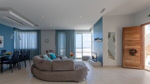 A modern living room with a curved sofa, a dining area with chairs, a staircase, and a large window with curtains. - Sea-View Apartment at WatersEdge Complex with two pools (Marsaskala)