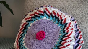 A crocheted pillow with a central flower design, placed on a chair with a textured cushion.