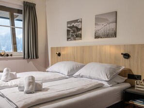 A hotel room with a wooden headboard, two white pillows, a neatly made bed with towels, a window with curtains, and two framed pictures on the wall. - Hilltop Apartment in Viehhofen With hot tub (Viehhofen)
