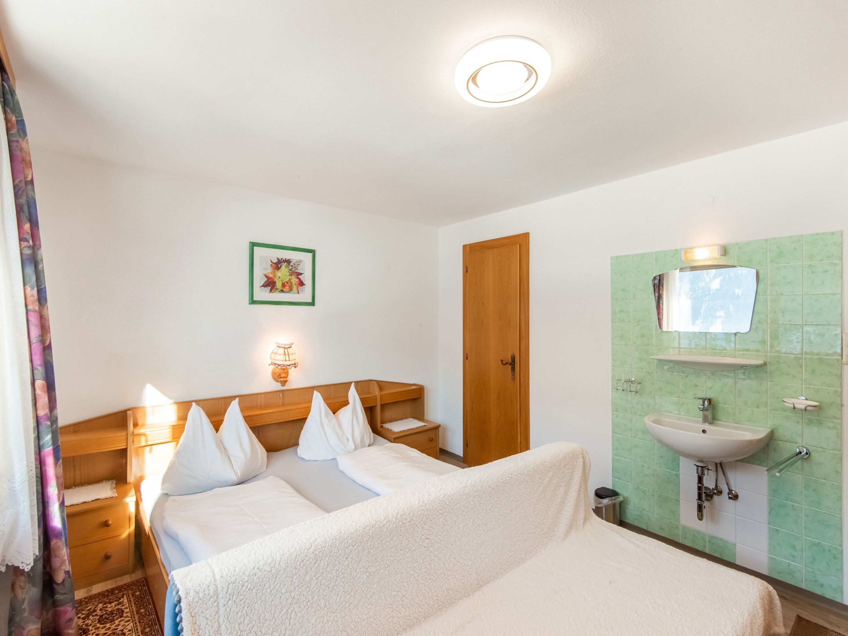 Un dormitorio con cama, mesitas de noche, lámpara de pared, una puerta y baño con lavabo y espejo.