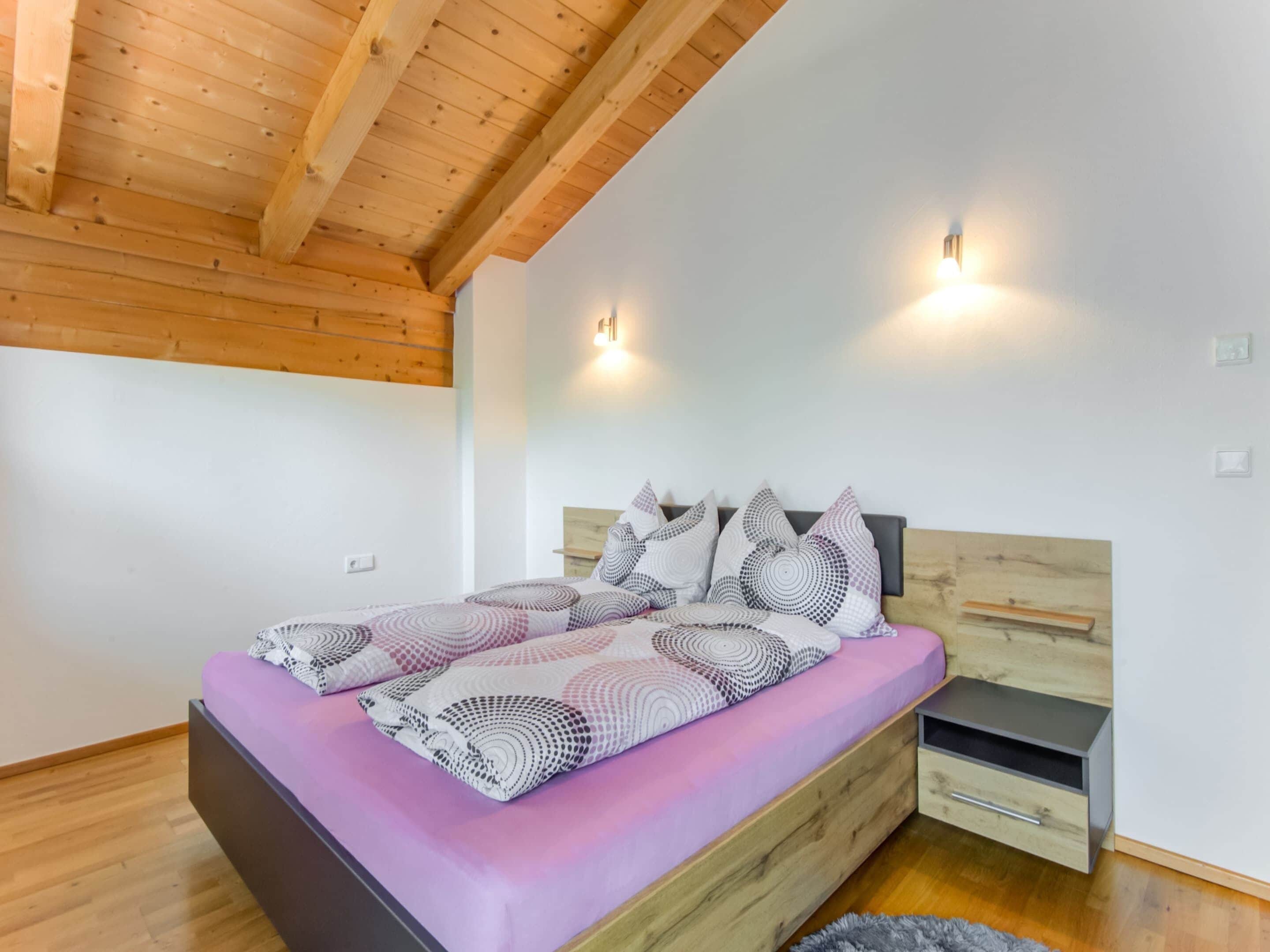 A bedroom with a wooden ceiling, a bed with patterned bedding, two wall-mounted lights, and a nightstand with a drawer.