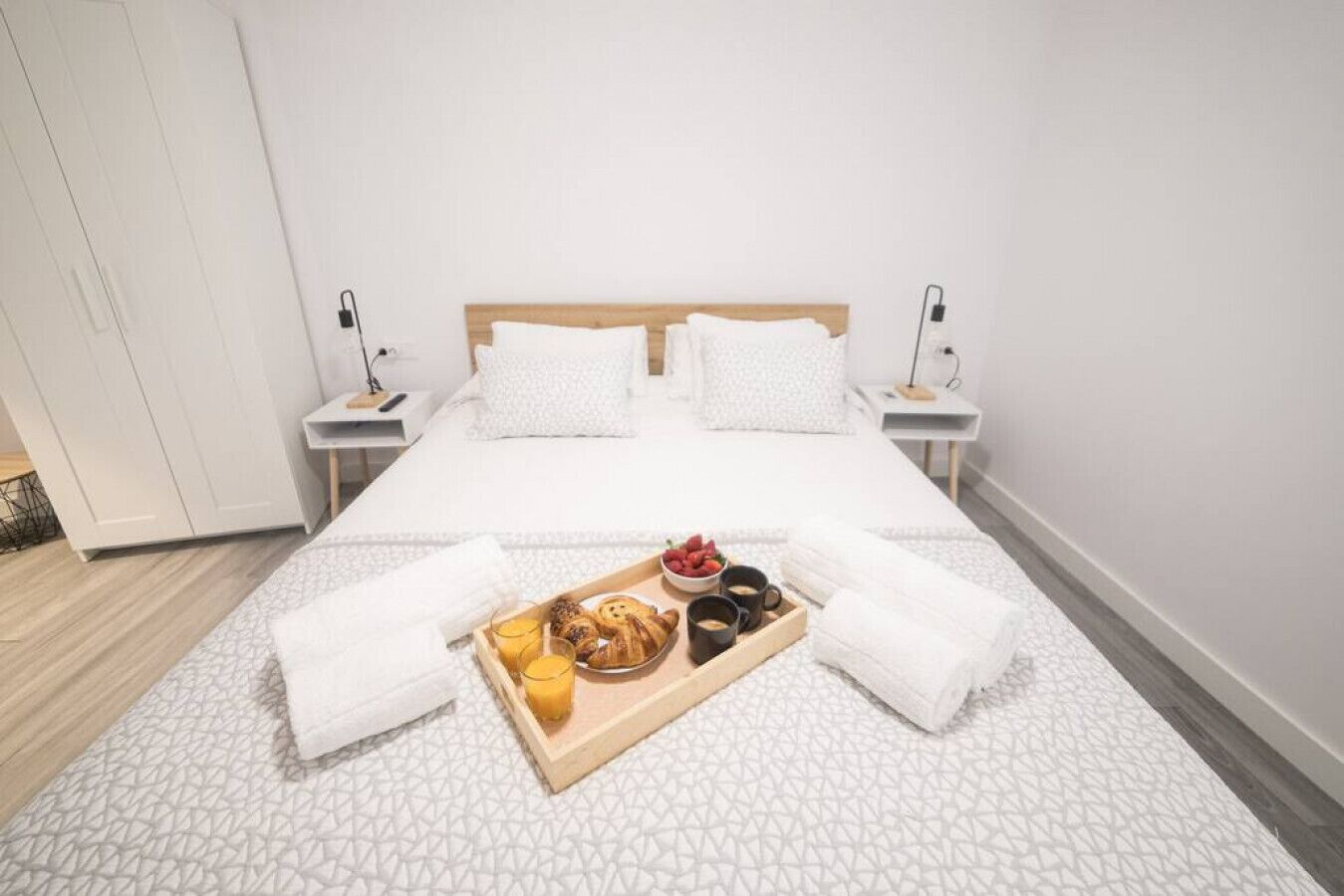 A neatly made bed with white linens, a tray of breakfast items including croissants, fruit, and coffee, and two bedside tables with lamps.