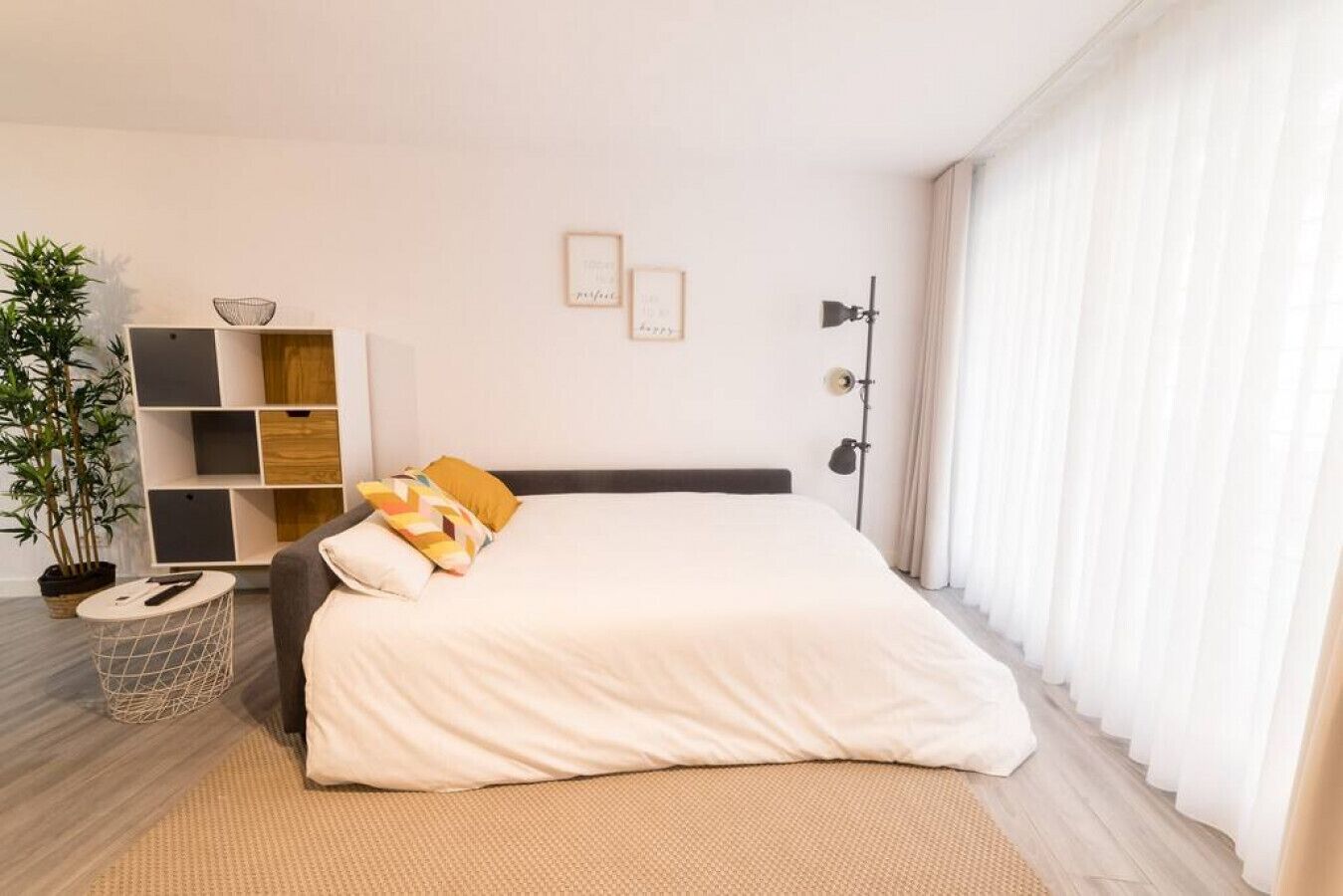 A modern bedroom with a large bed, a side table, a plant, and a bookshelf.