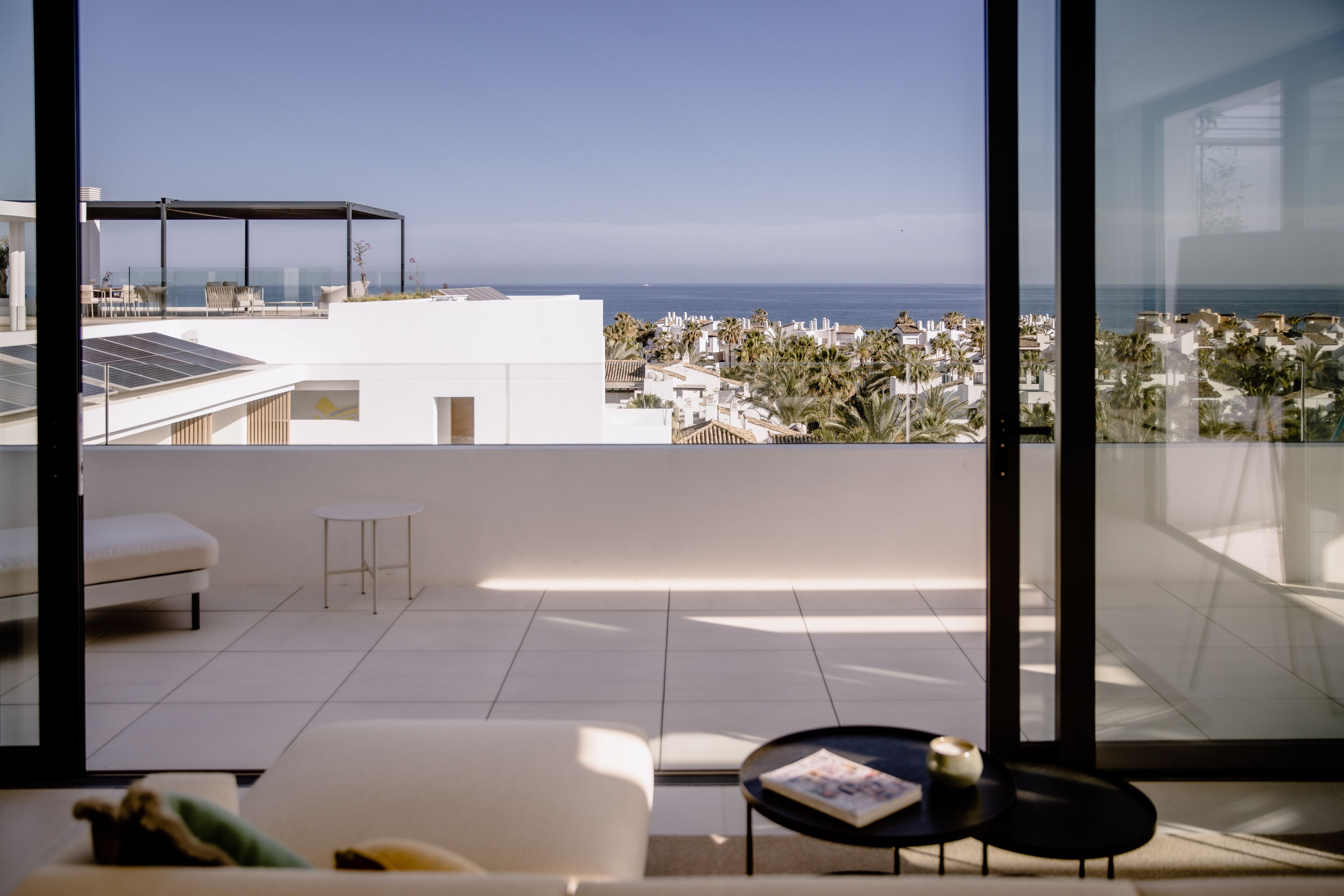 A modern room with a large window overlooking a coastal view, featuring a sofa, a small table, and a glass railing.