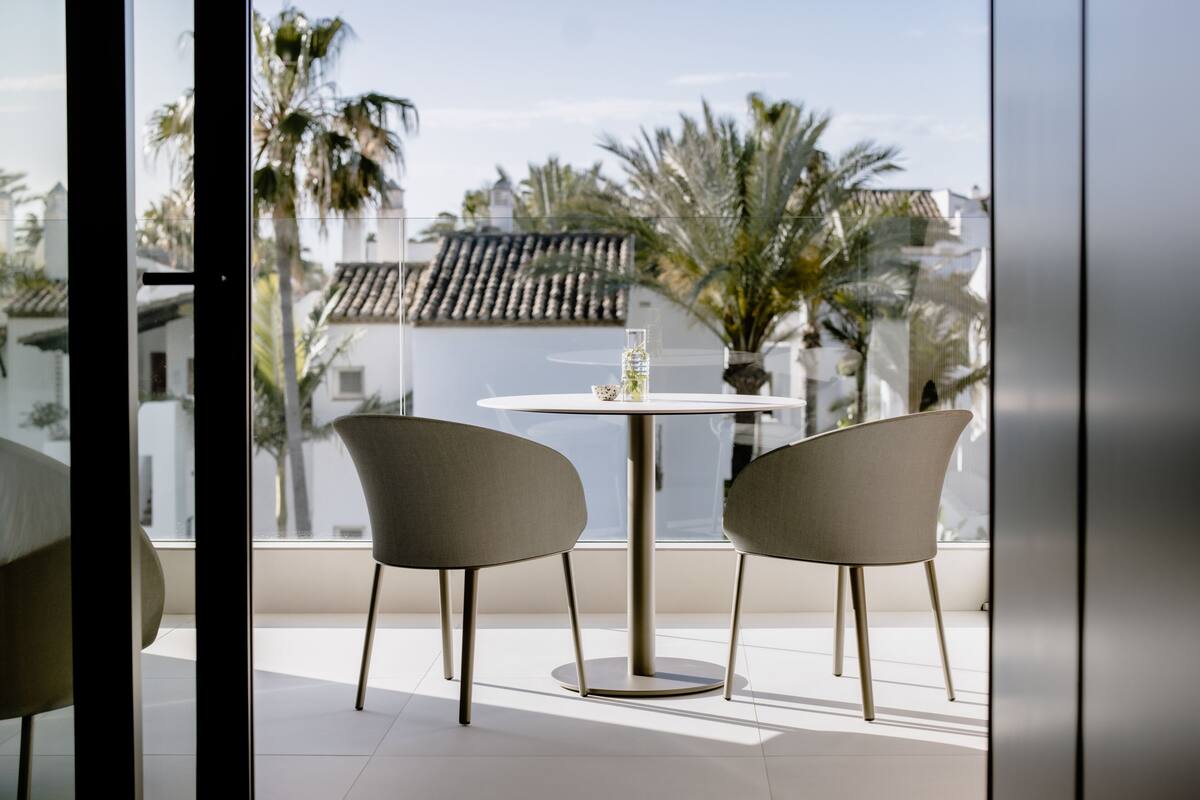 A balcony with two chairs and a table, overlooking a residential area with palm trees and houses.